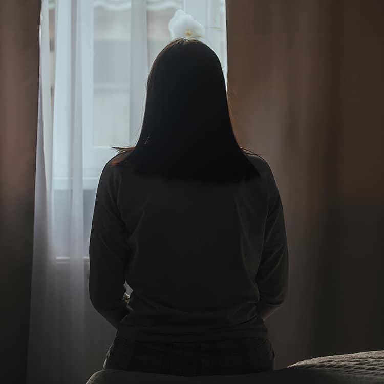 Silhouette of a young woman with ME, in bed, against the background of a window in a darkened room. / Silhouette d'une jeune femme avec l'EM, au lit, avec pour fond une fenêtre dans une pièce sombre.